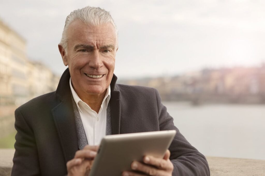pexels photo 3782213 Elderly man smiling while using a tablet outdoors by a riverside.